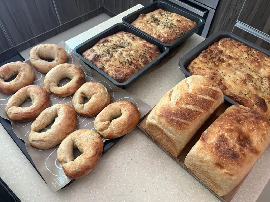 Homemade sourdough bagels, focaccia and bread loaves baked in loaf pans on a kitchen counter.