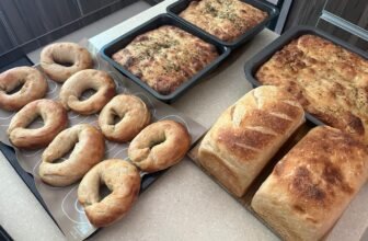 Homemade sourdough bread, golden focaccia baked in loaf pans, and freshly baked bagels arranged on a kitchen counter.